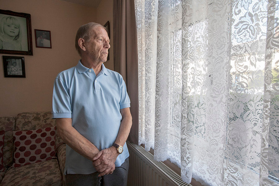 Man standing by a window with lace curtains in a room with framed pictures on the wall.