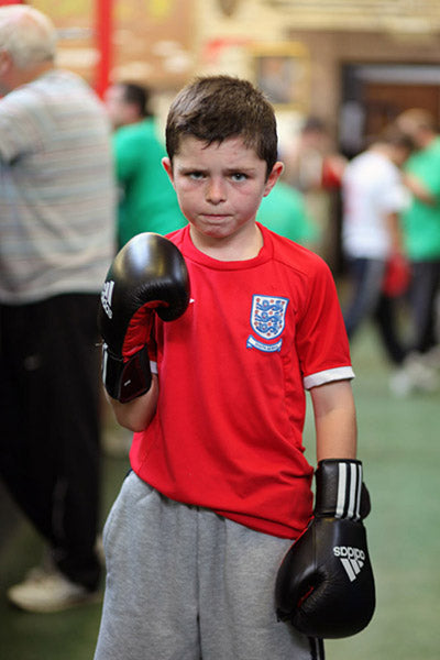 Child wearing boxing gloves and a red shirt with a logo, standing in a gymnasium.