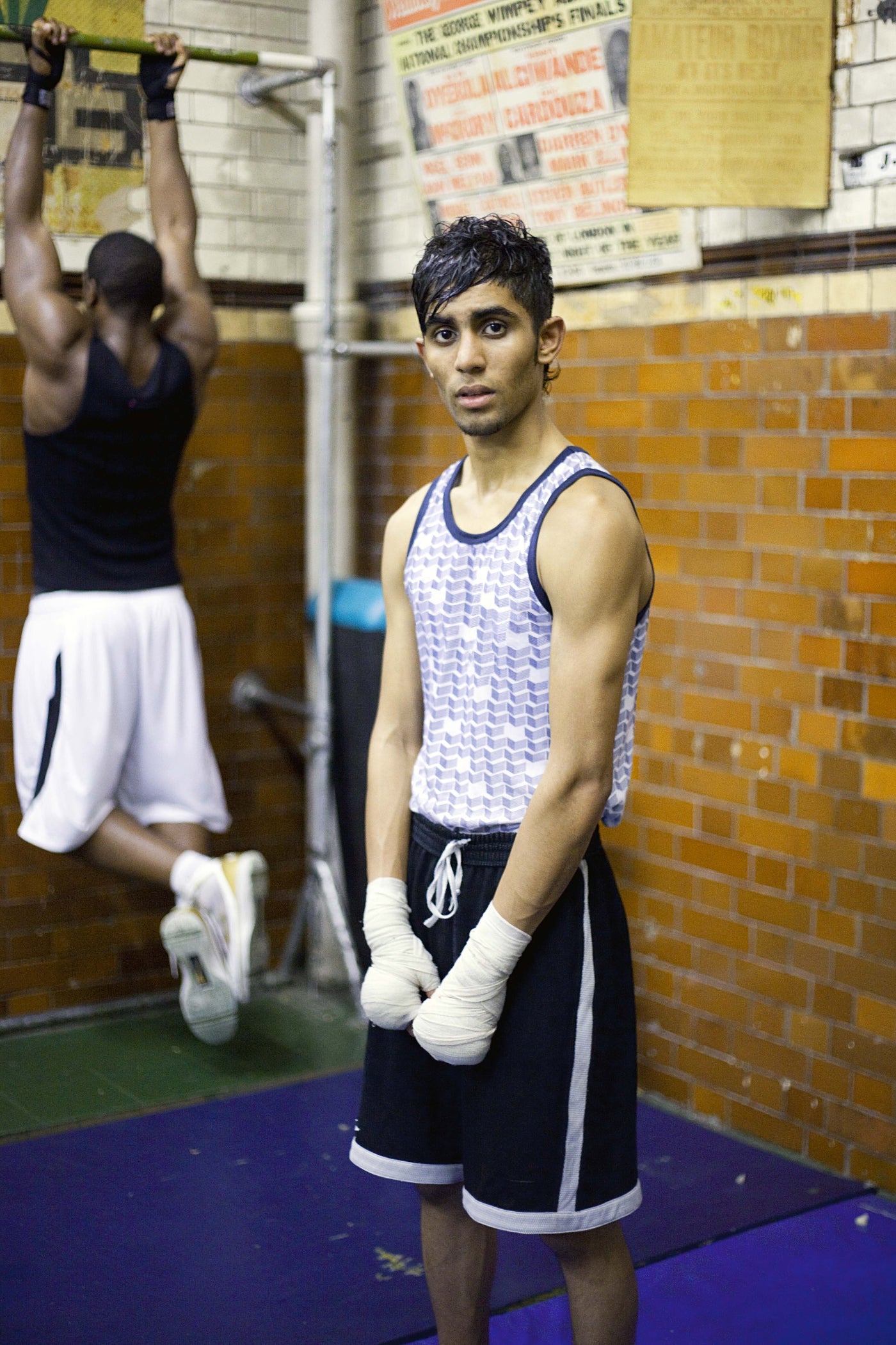 Person in a gym wearing boxing gloves and shorts, with another person in the background.