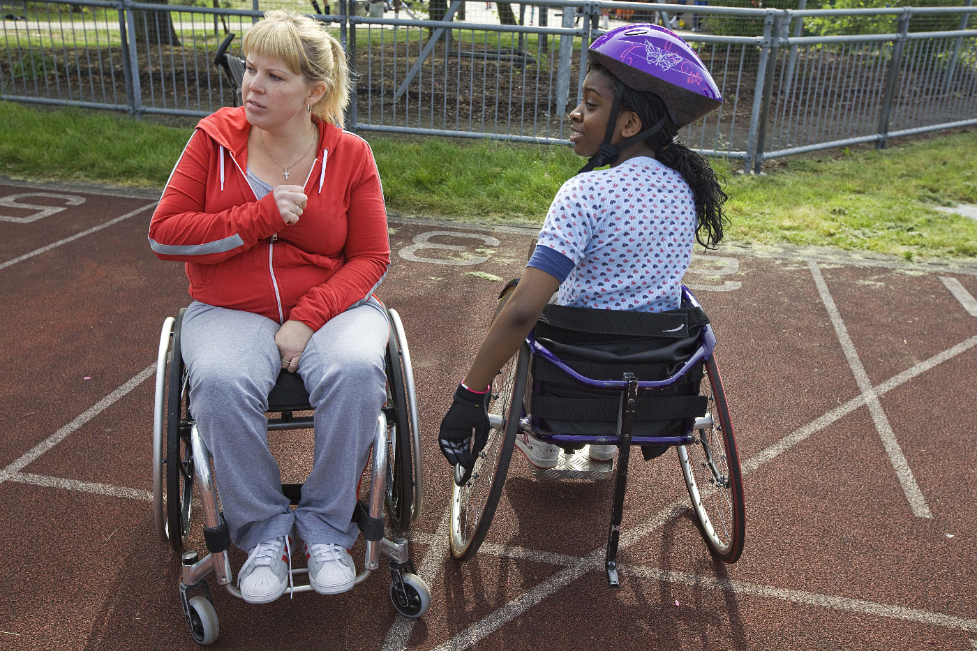 Two women in wheelchairs on a track, one wearing a red jacket and the other in a purple helmet.