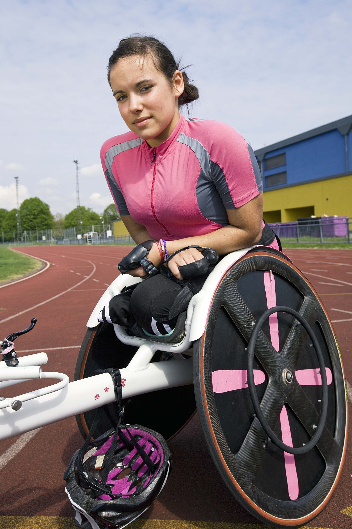 Person in a pink and gray cycling outfit sitting on a racing wheelchair on a track.