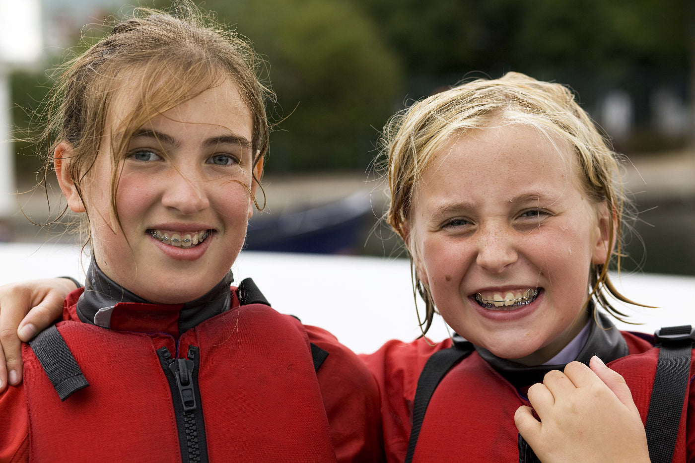 Two young girls in red life jackets smiling outdoors with wet hair. 