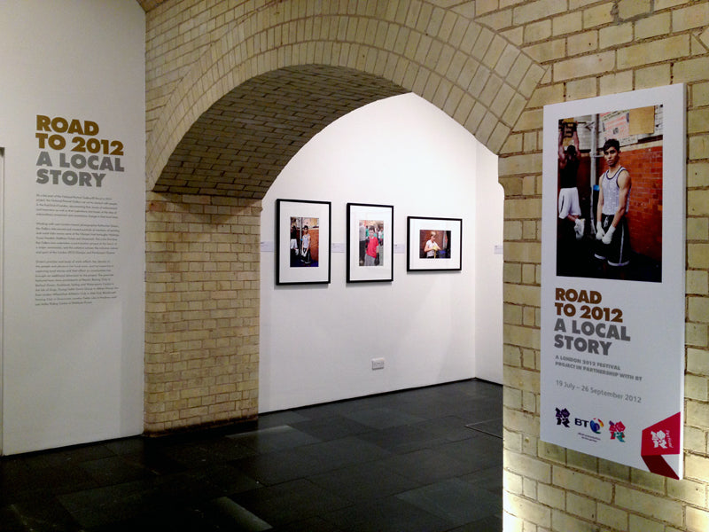 Exhibition space with framed pictures and a large poster on a brick wall.