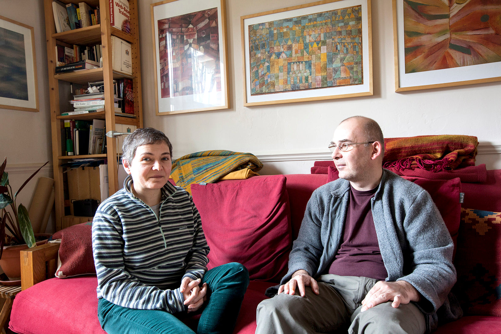 Two people sitting on a couch in a living room with books and artwork on shelves.
