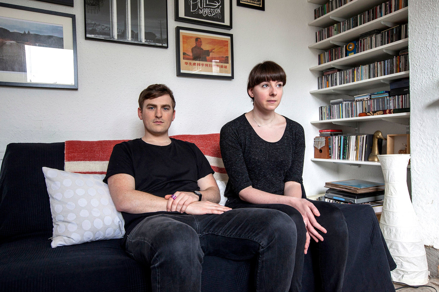 Two people sitting on a couch in a room with a bookshelf and framed pictures.