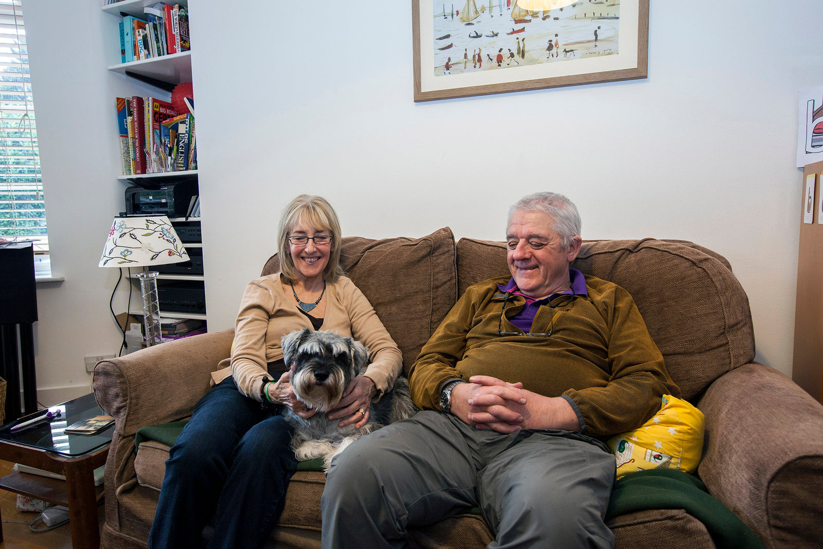 Two people sitting on a couch with a small dog, in a living room setting.