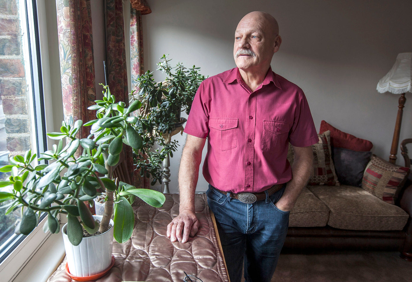 Man in a pink shirt standing in a living room with plants and a couch.