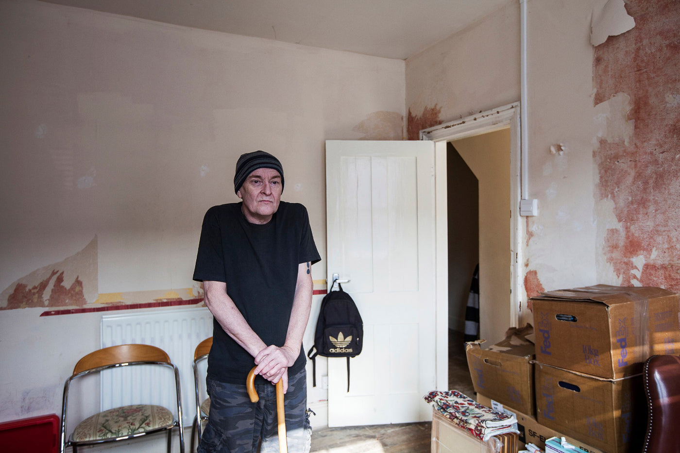 Person standing in a room with peeling walls and furniture, including a chair and a backpack.