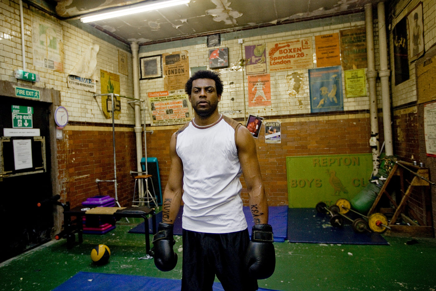 Man in a white tank top and black boxing gloves standing in a boxing gym with posters on the wall.