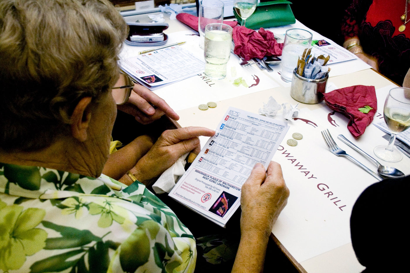 Person looking at a list of greyhounds to bet on at a restaurant table with various items on it.