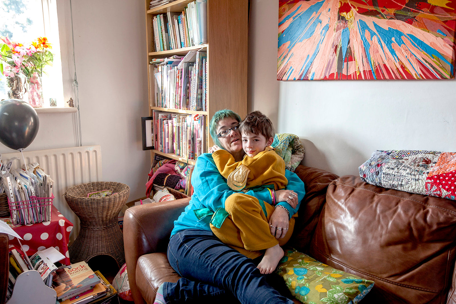 Woman and child sitting on a brown sofa in a cozy living room with books and a colorful painting.