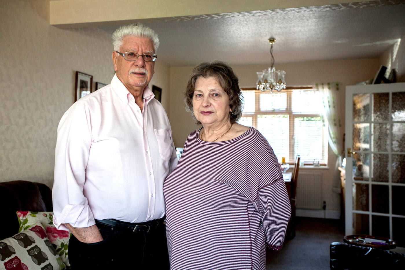Two elderly people standing in a living room with a chandelier and window in the background.
