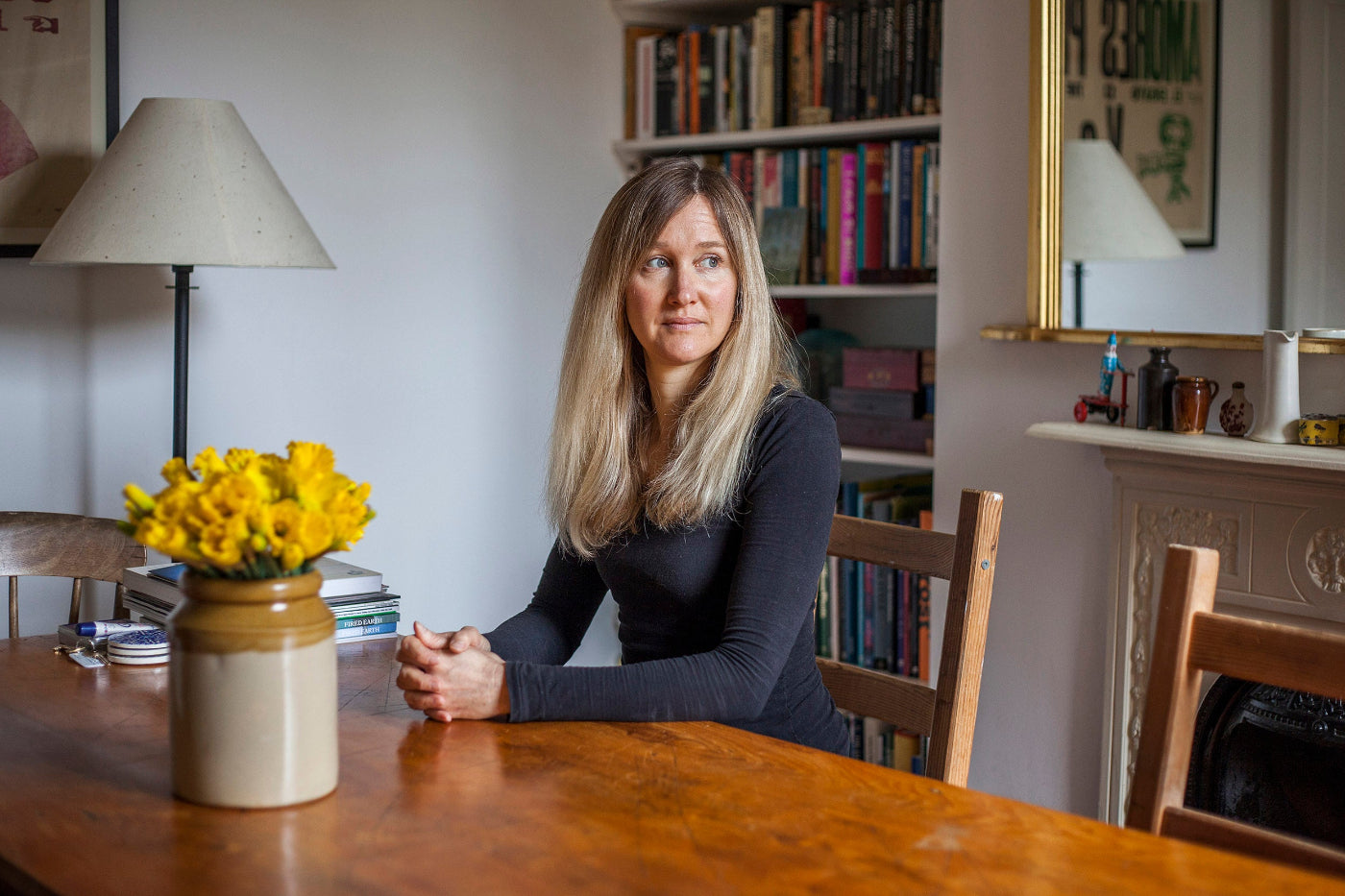 Woman sitting at a wooden table with a vase of yellow flowers in a room with bookshelves and a lamp.