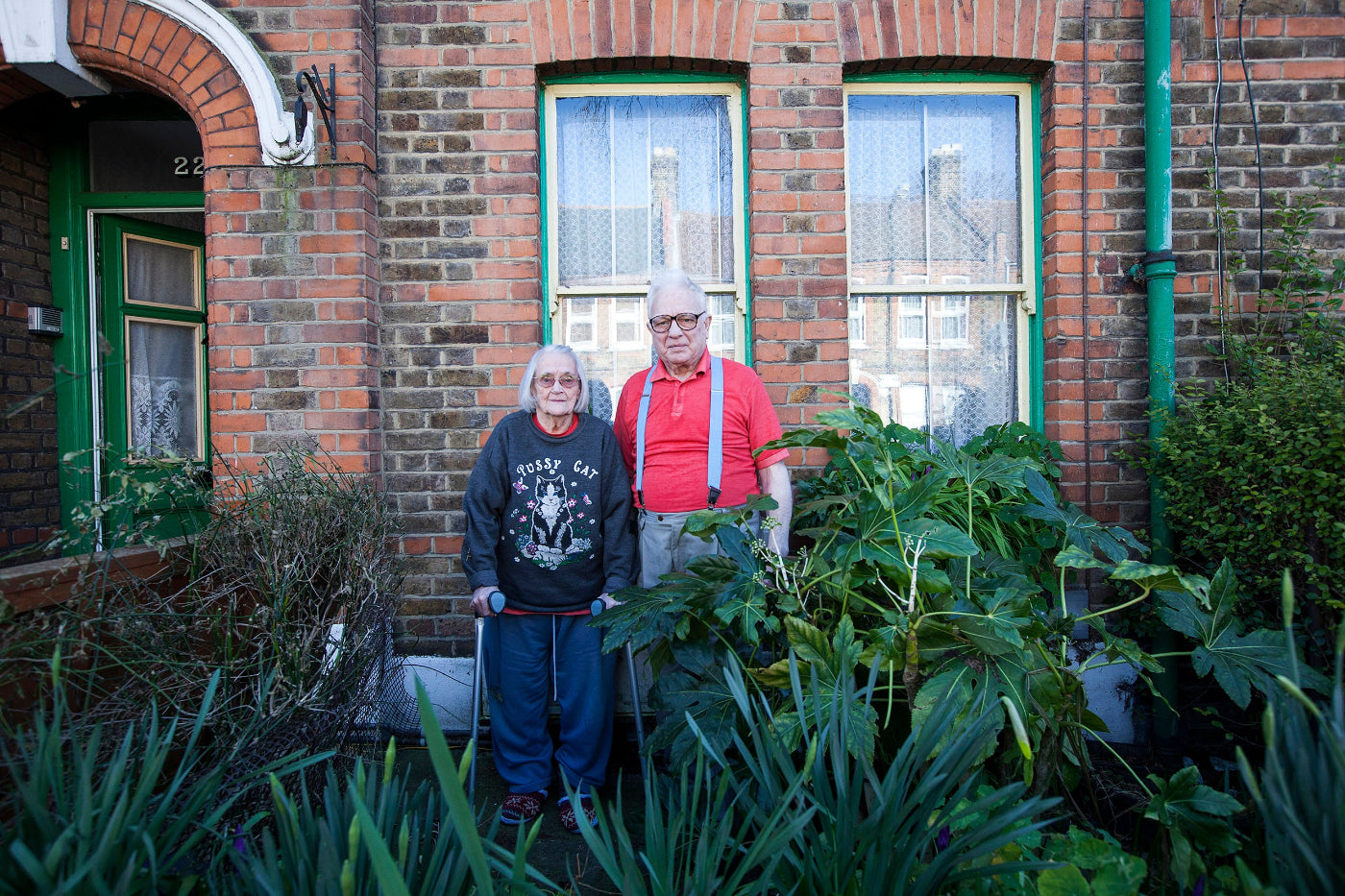 Two people standing in front of a brick building with plants around them.
