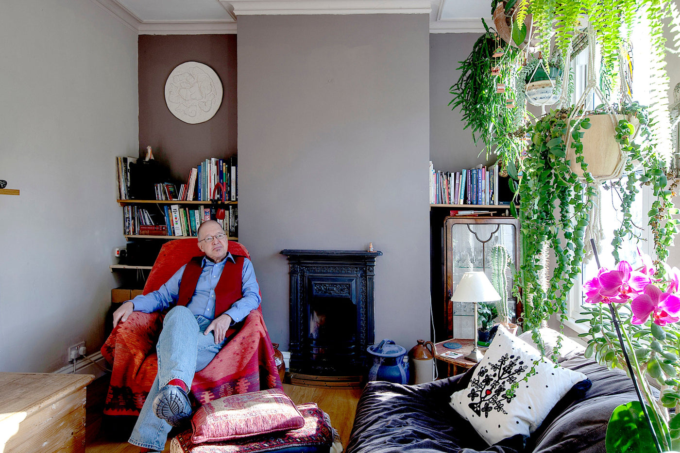 Man sitting in a cozy living room with books, a fireplace, and plants.