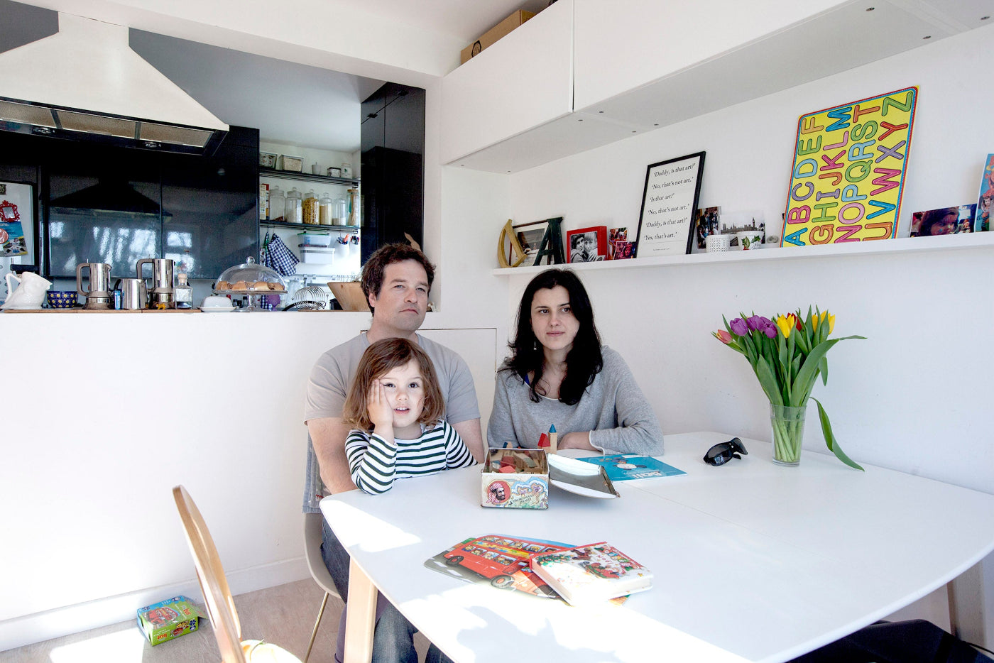Family of three sitting at a kitchen table with toys and books, surrounded by modern kitchen appliances and decor.