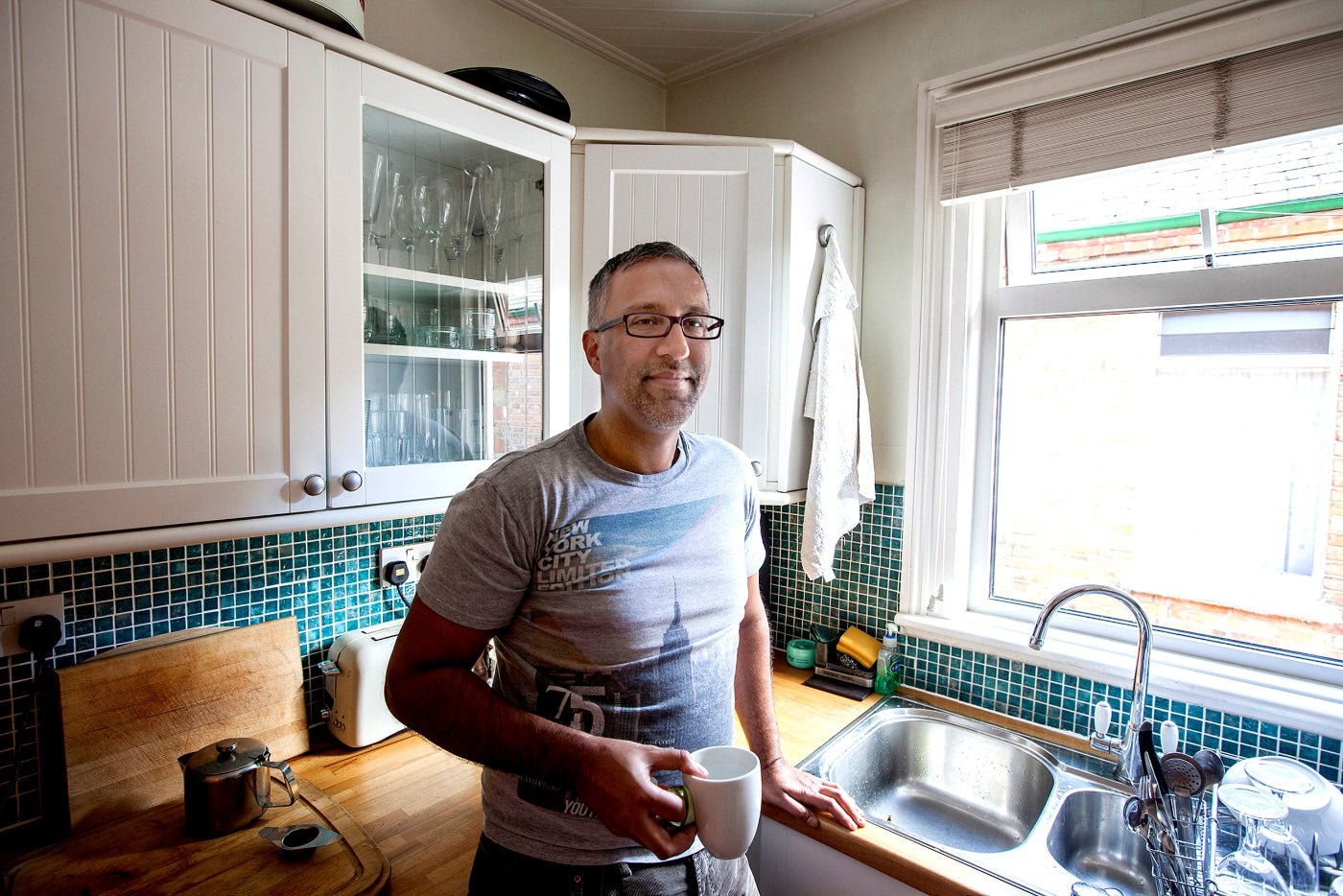 Man standing in a kitchen holding a mug, with cabinets and a sink visible.