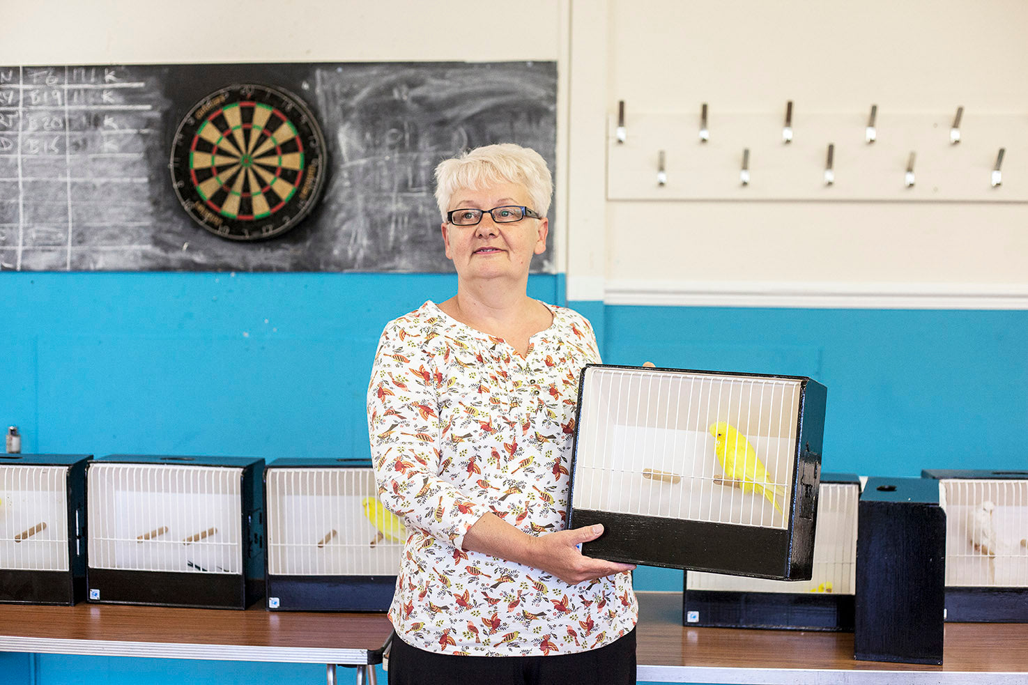 Person holding a cage with a yellow bird inside, in front of a display of other birds in cages on a table